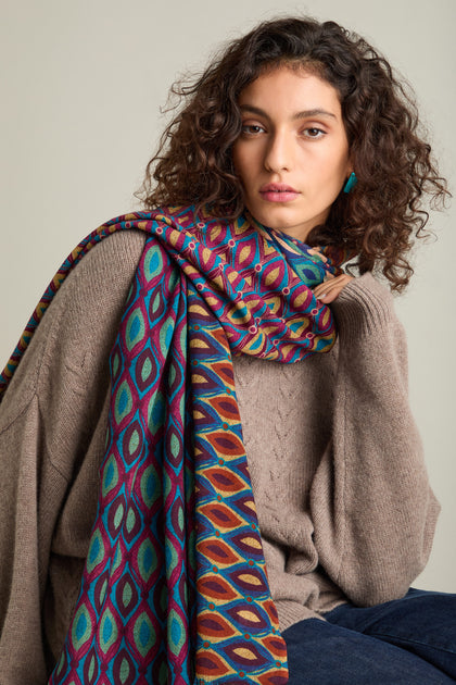 Person with curly hair wearing a beige sweater and a vibrant retro wool twill scarf, gazing towards the camera.