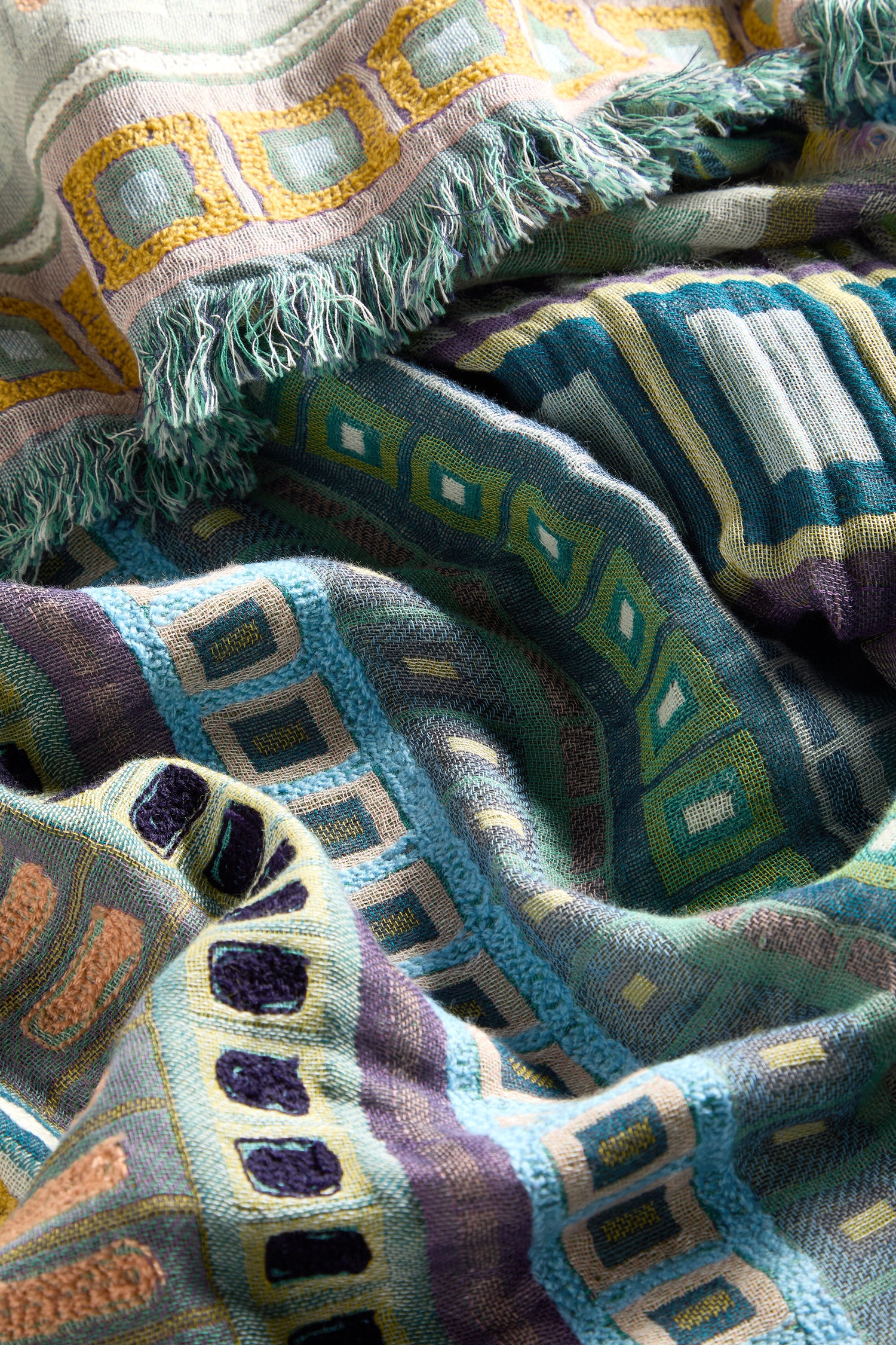 Brightly woven textile boasting geometric patterns and fringed edges. The Textured Cotton Scarf, ideal for seasonal transitions, showcases a vibrant array of blue, green, purple, and yellow squares and rectangles.