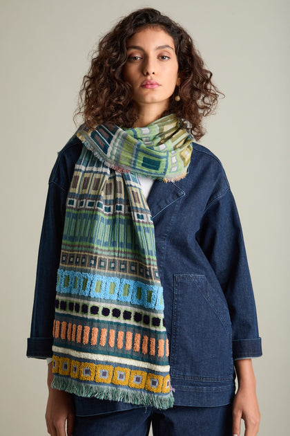 A person with curly hair wearing a dark blue denim outfit and the Textured Cotton Scarf, featuring vibrant colors and rich textures, stands against a plain background, making it an ideal look for transitioning between seasons.