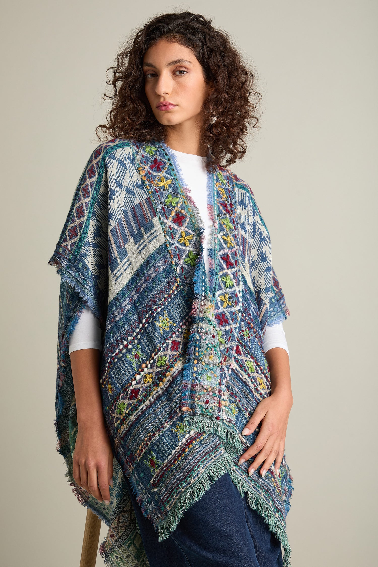 A person stands against a plain background wearing a colorful, patterned poncho with fringe, paired with a white shirt and blue jeans. They have curly hair and the Embroidered Cotton Shawl draped elegantly over one shoulder, adding a touch of luxurious accessory to their ensemble.