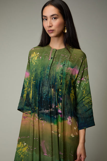 A woman with long dark hair wears the Monet Pleated Dress, featuring a green abstract print and pleated details, paired with gold earrings against a neutral background.