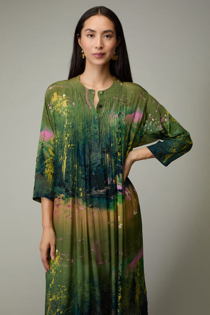 A woman in the Monet Pleated Dress, a long green piece with three-quarter sleeves and an abstract print, stands against a plain background facing the camera with a neutral expression.