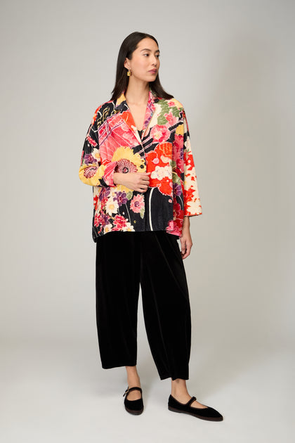A woman stands in a studio wearing the Abstract Poppy Velvet Jacket—wearable art—paired with black wide-leg pants and black flat shoes, gazing slightly to the side.