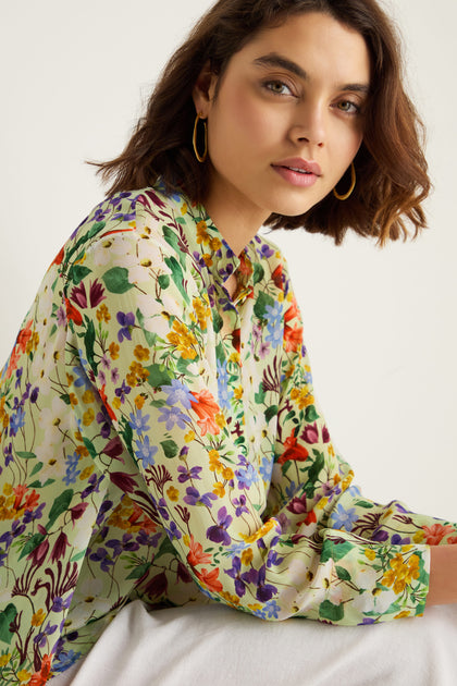 A woman with short brown hair, wearing the Country Garden Cupro Shirt—a colorful, long-sleeve floral blouse—and gold hoop earrings sits against a neutral background.