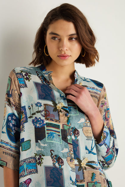 A woman with short brown hair models the Postcard Print Cupro Shirt, which features stamp and illustration designs. She stands against a white background, looking slightly to the side.