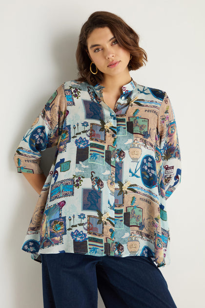 A woman models the Postcard Print Cupro Shirt with blue and brown prints, styled with dark pants, standing against a plain light backdrop.