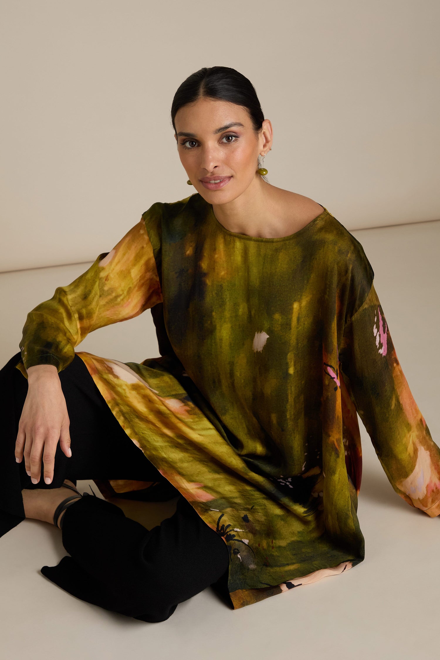 A seated person wearing a long-sleeved, Autumnal Watercolour Tunic and black pants, posing against a beige background, showcases a relaxed silhouette perfect for any transitional wardrobe.