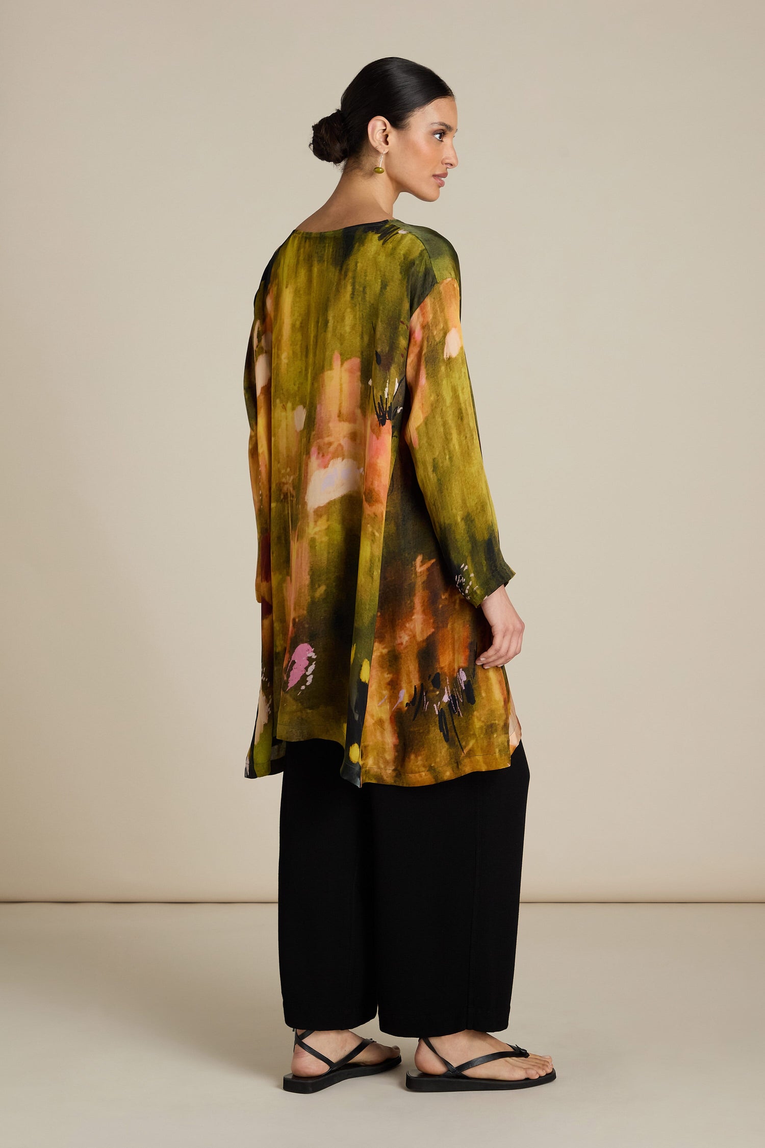 A person stands facing slightly away, wearing a long, multicolored Autumnal Watercolour Tunic with black pants and black sandals. The Autumnal Watercolour Tunic features abstract patterns in green, yellow, and pink tones, perfect for a transitional wardrobe with its relaxed silhouette.