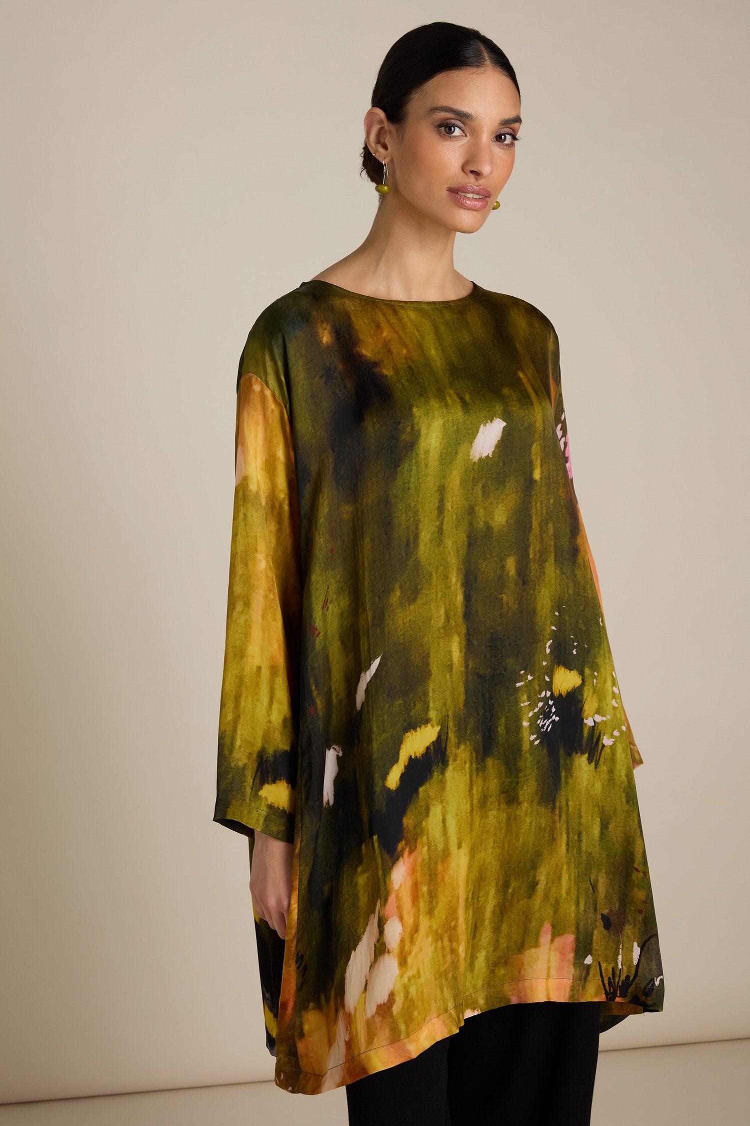 A woman is standing against a plain background, wearing a loose-fitting, green and yellow Autumnal Watercolour Tunic with long sleeves. Featuring a relaxed silhouette, her dark hair is pulled back as she looks directly at the camera. Perfect for a transitional wardrobe.