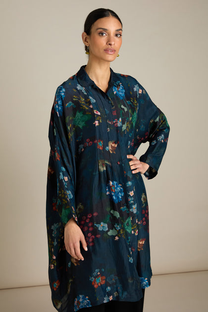 The original sentence already uses the product name "Monet Button Through Dress," so no replacement is necessary. The sentence is:

A woman stands with one hand on her hip, wearing a long, dark Monet Button Through Dress adorned with floral patterns. Her dark hair is tied back, and she wears earrings. The background is plain and light-colored, creating an elegant depiction reminiscent of impressionist art.