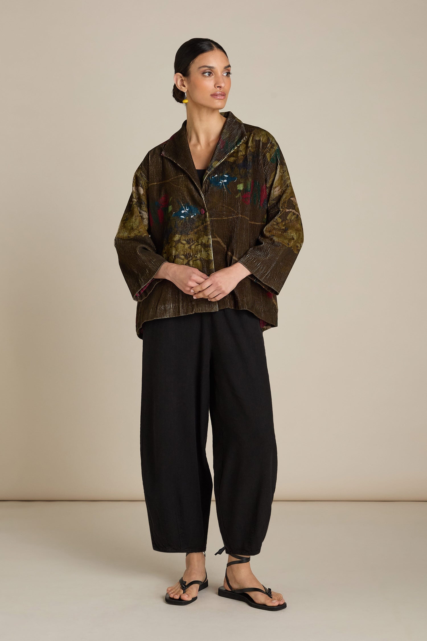 A woman stands against a plain backdrop, wearing a loose-fitting, Cord Printed Jacket with one-button fastening, black top, black pants, and sandals. She has her hands lightly clasped in front.