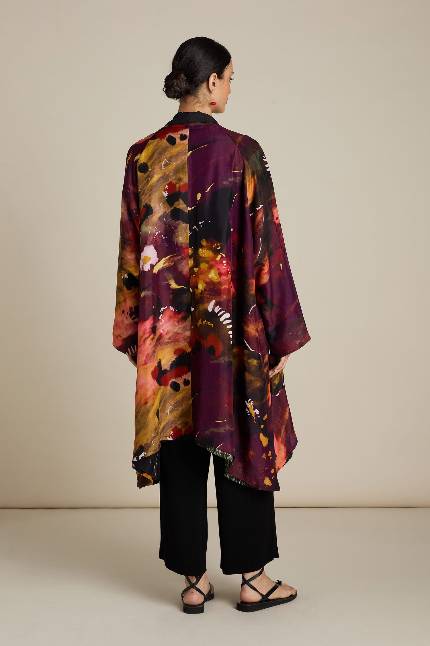 A person stands facing away, wearing a long, colorful Reversible Abstract Flower Print Jacket that exudes artful elegance. Paired with black pants and black sandals, the ensemble presents a relaxed silhouette.