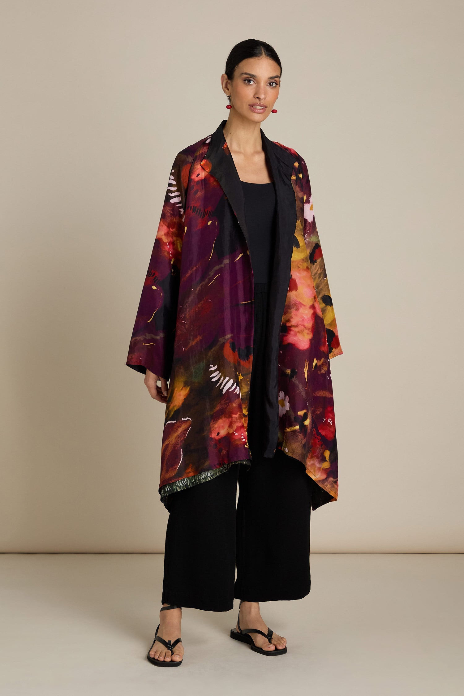 A person stands against a plain background wearing a Reversible Abstract Flower Print Jacket over a black inner outfit, including wide-legged pants and flat sandals. The relaxed silhouette and artful elegance of the ensemble create an effortlessly chic look.