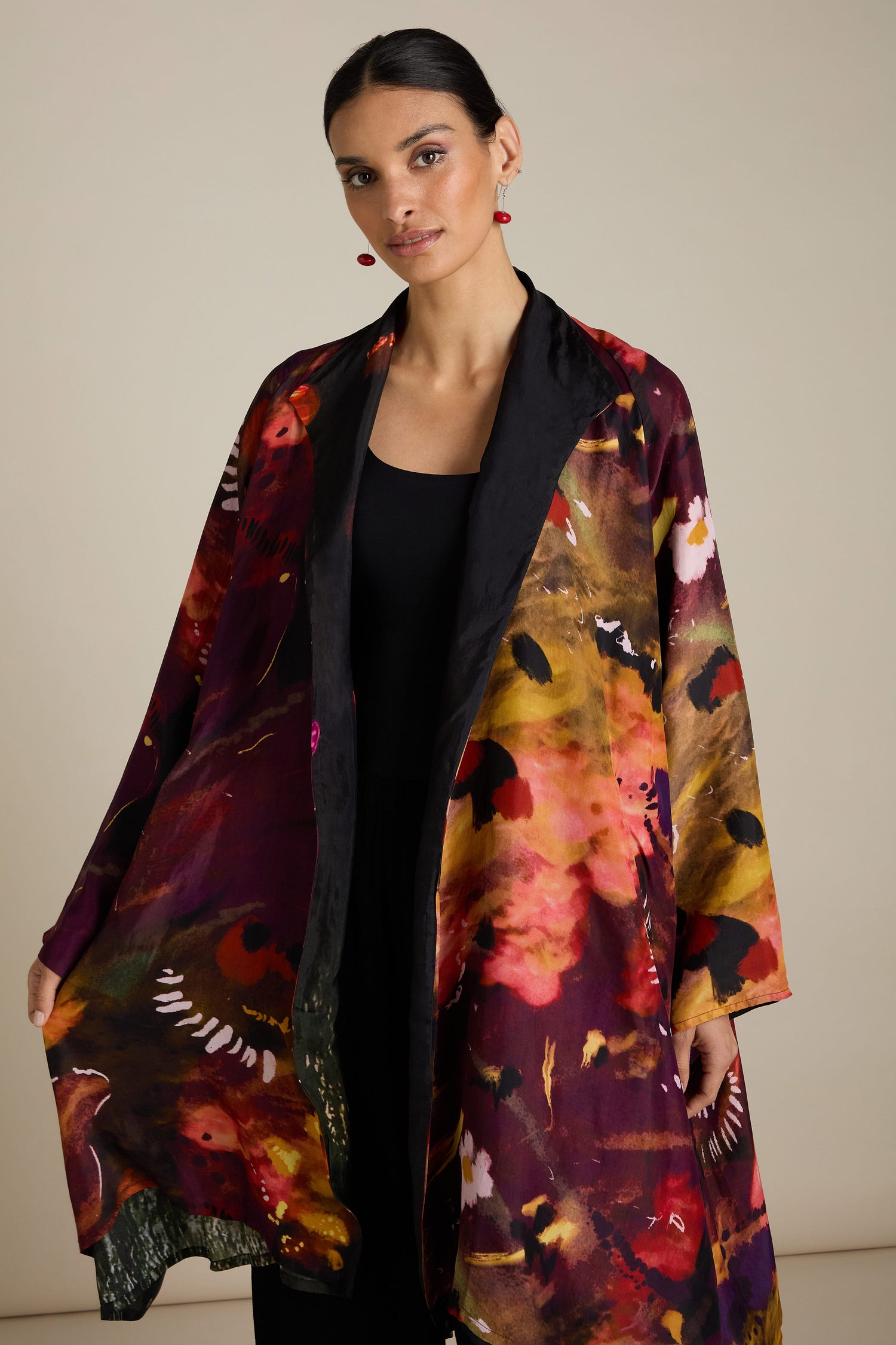 A person is wearing a colorful, patterned Reversible Abstract Flower Print Jacket with dark hues and floral designs, paired with a black outfit underneath. They have dark, tied-back hair and red earrings, embodying artful elegance with their relaxed silhouette.