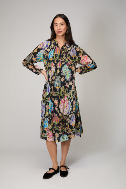 A woman in a studio wears the Protea Flower Dress—a striking black, knee-length piece adorned with vibrant florals and paisley designs—styled with black flats.