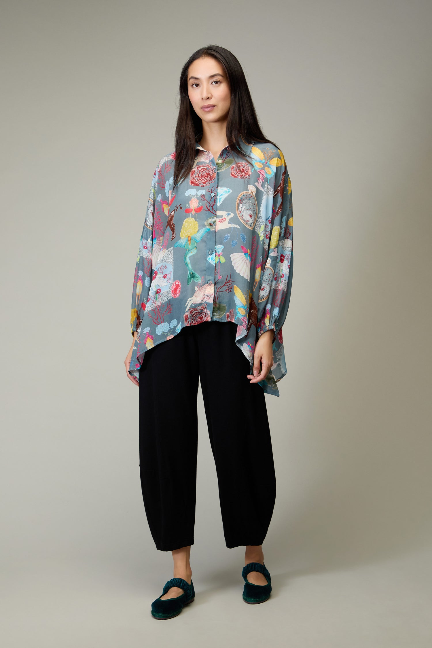 A woman stands against a plain background wearing the Fantasy Print Shirt, featuring a colorful collage in the Yavi illustrative style, paired with black pants and dark shoes.