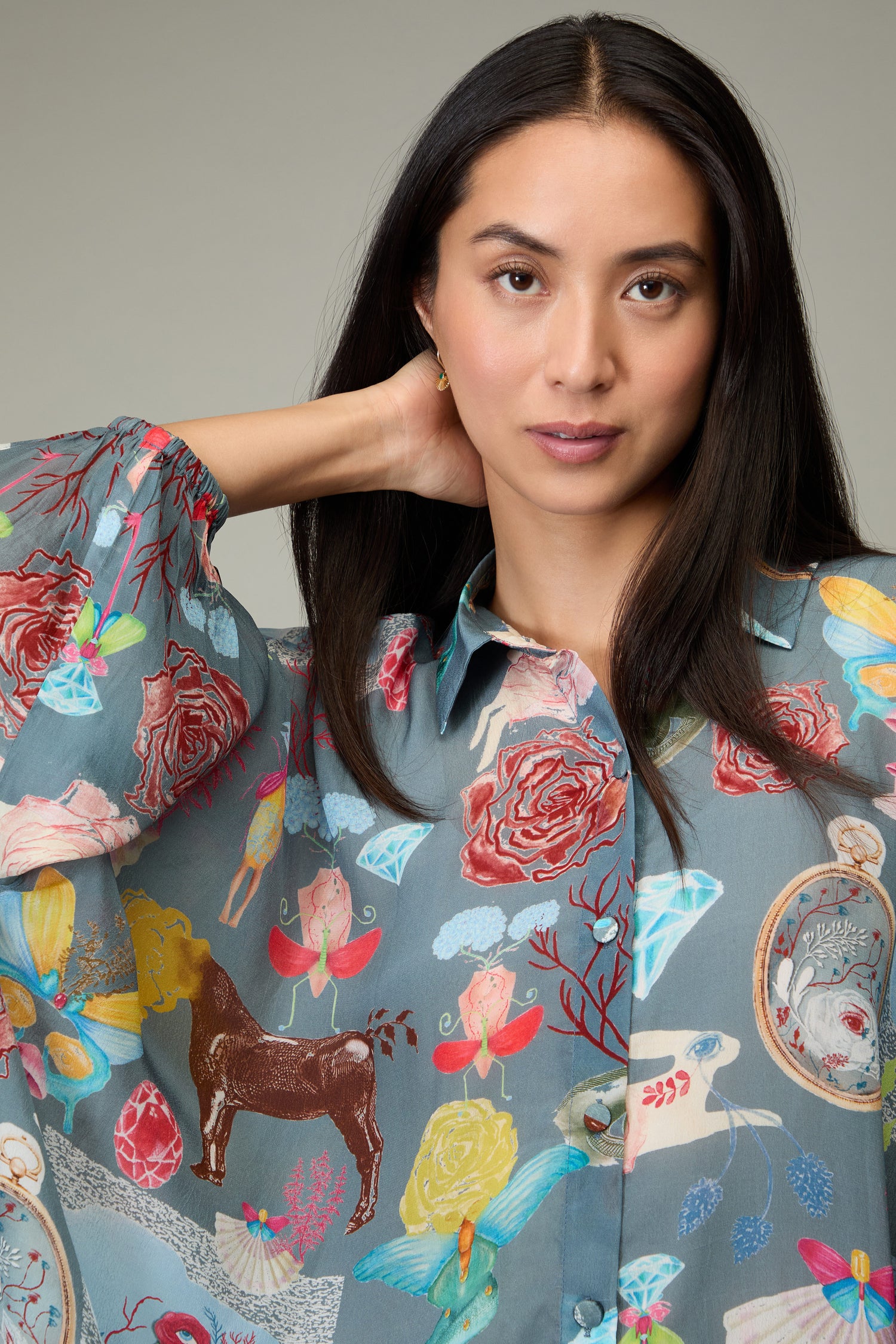 A woman with long dark hair wears the Fantasy Print Shirt in gray, featuring colorful illustrations of flowers, horses, birds, and abstract patterns—a true showcase of Yavi's distinctive illustrative style.