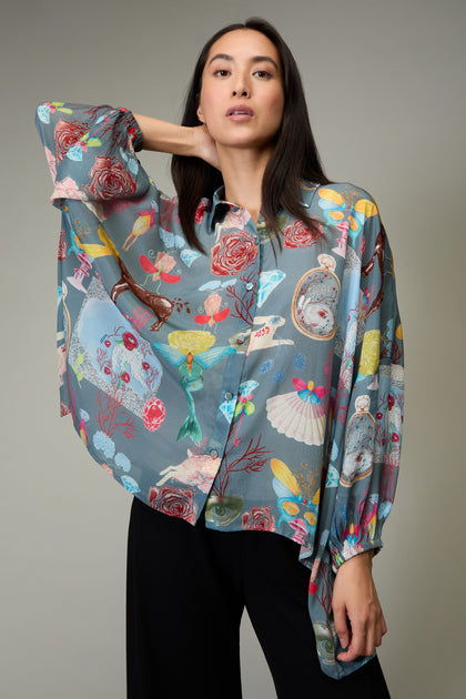 A woman in black pants wears the Fantasy Print Shirt, featuring a whimsical collage of flowers, birds, and objects in Yavi's illustrative style. She stands against a plain gray background.