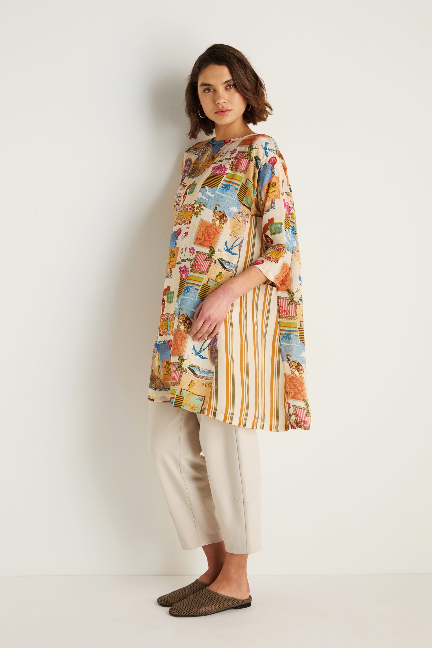 A woman stands against a white wall wearing the Postcard Print Cupro Tunic with hand-drawn prints and stripes, paired with beige cropped pants and brown flat shoes.