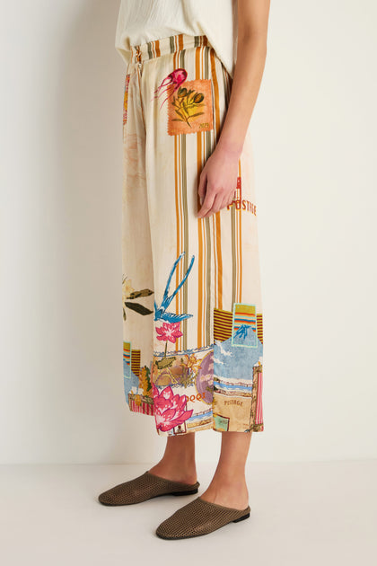 A person stands against a white wall wearing Postcard Print Stripe Trousers with wide legs and colorful patchwork inspired by Indian textile artistry, paired with brown woven slip-on shoes.