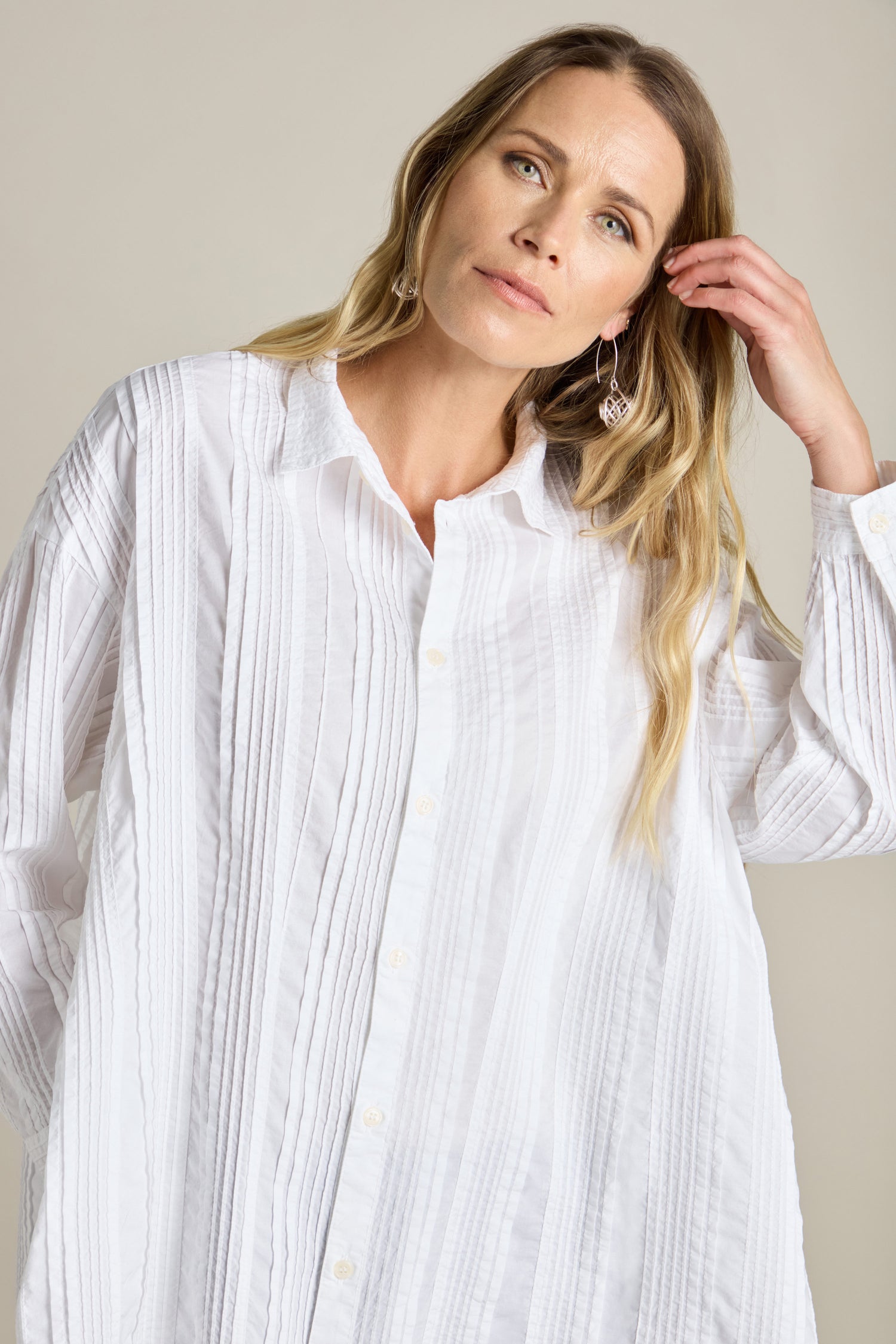 A woman with long blonde hair is wearing a white Multi Pleat Shirt with an oversized fit and silver earrings, touching her hair with her right hand. She has a neutral expression and is standing against a beige background. Her stylish attire resembles the unique designs of Yacco Maricard.