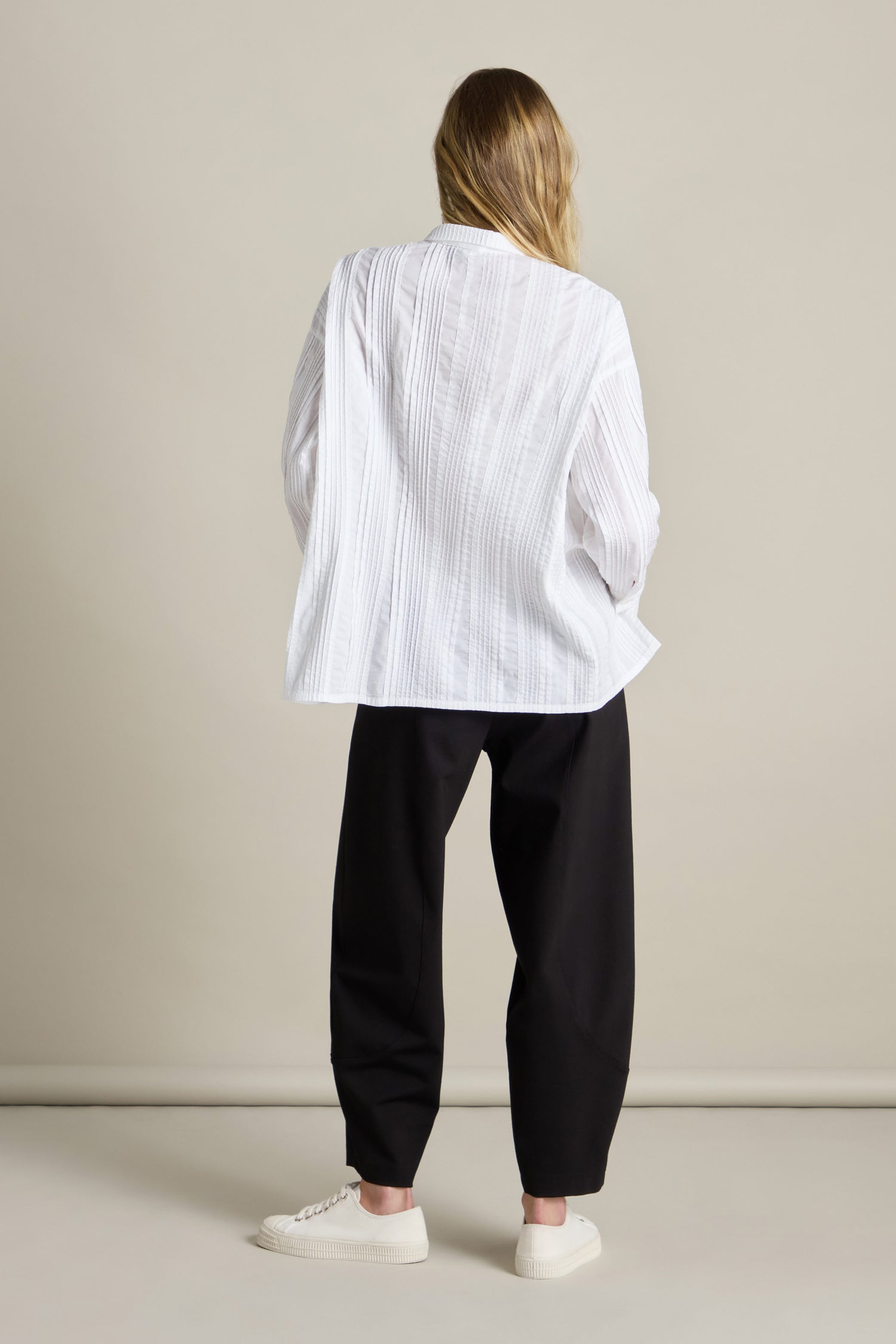 Person with long hair stands facing away, wearing a sheer white Multi Pleat Shirt with an oversized fit, loose black pants, and white sneakers against a plain background.