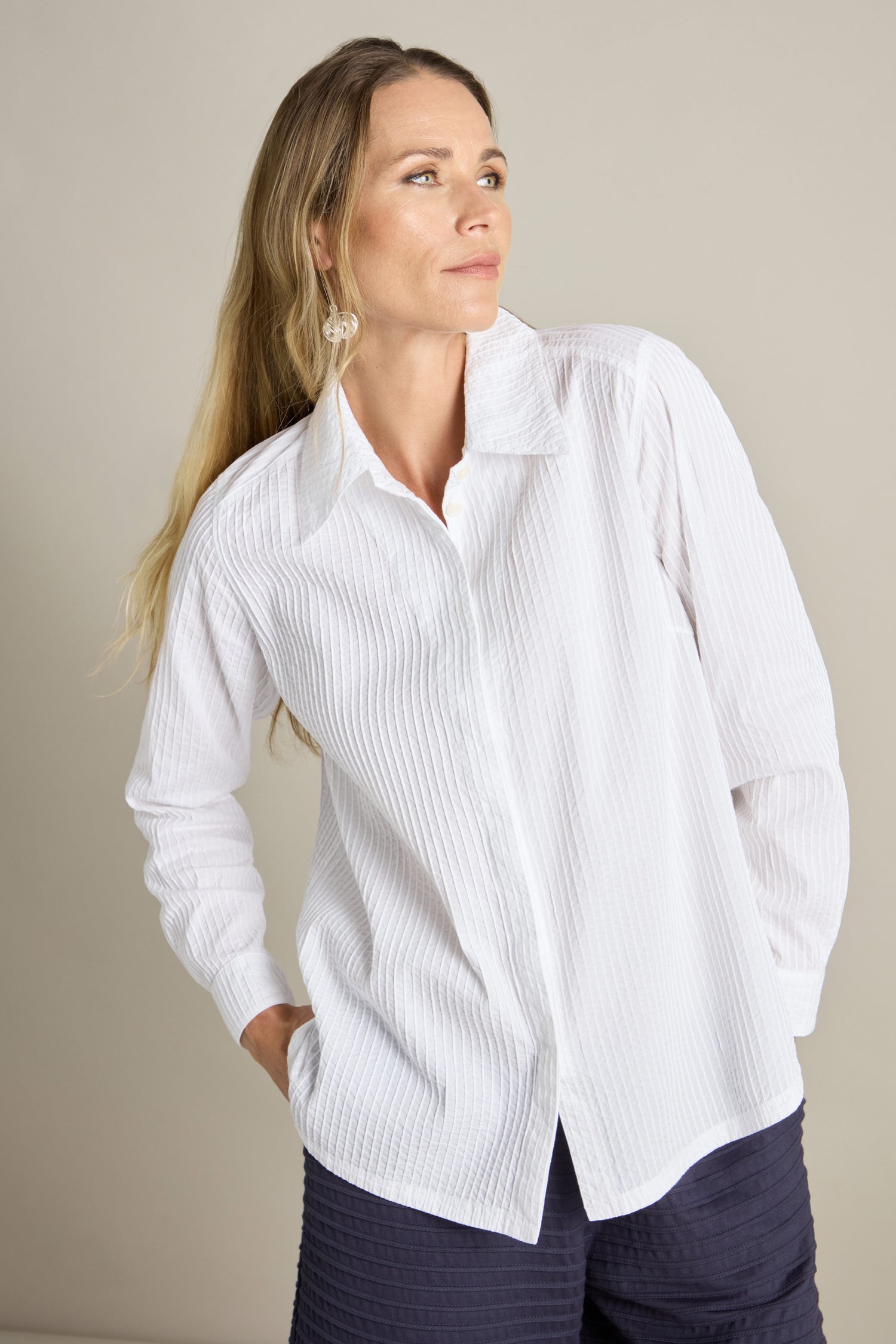 A person wearing Yacco Maricard's white Fitted Pleat Shirt and dark pants stands with hands in pockets, looking to the side, showcasing a tailored look.