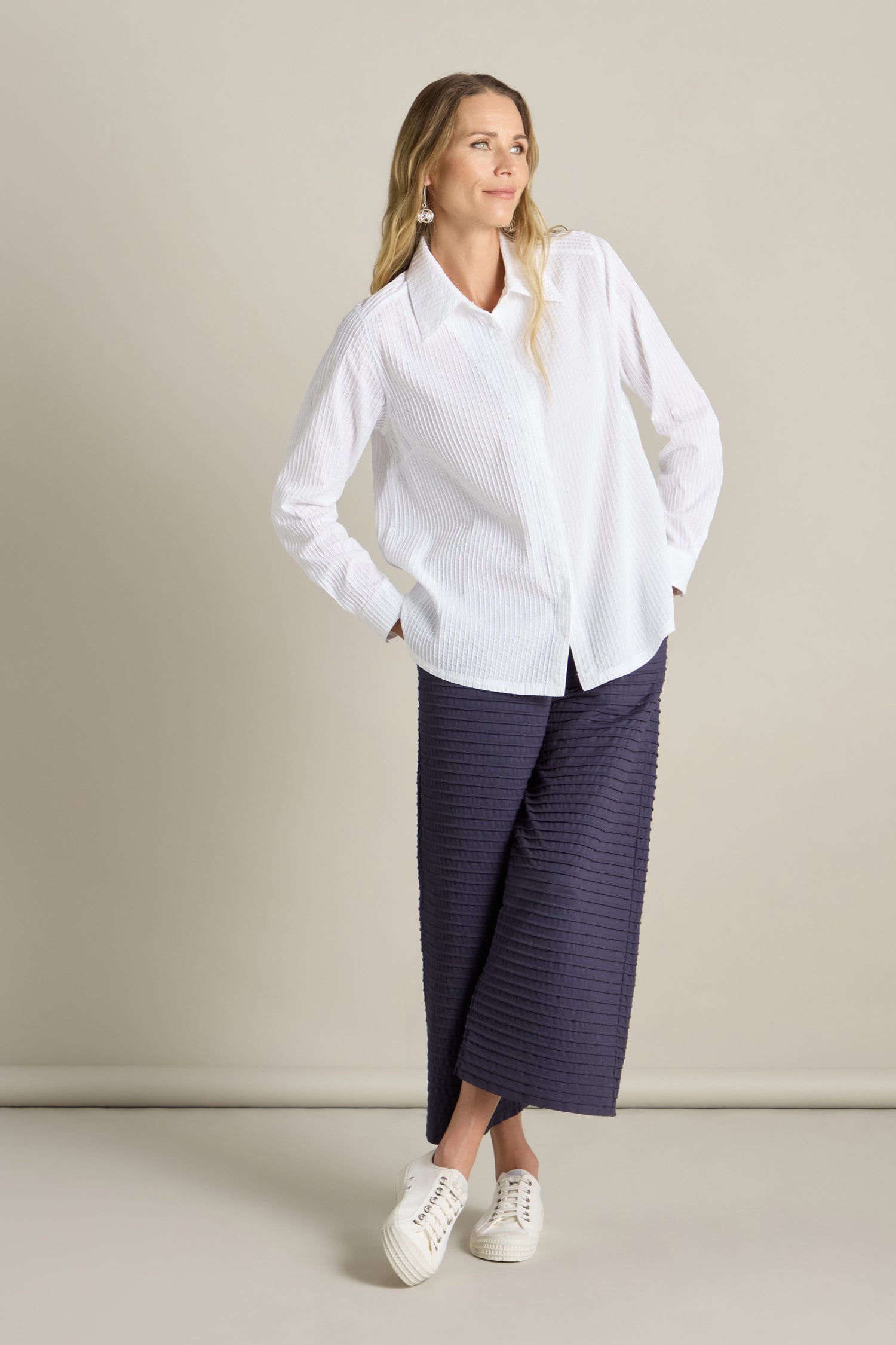 A woman stands against a plain background, wearing a Fitted Pleat Shirt, dark wide-legged pants, and white sneakers. She rests her hands in her pockets and looks off to the side, exuding a tailored look.