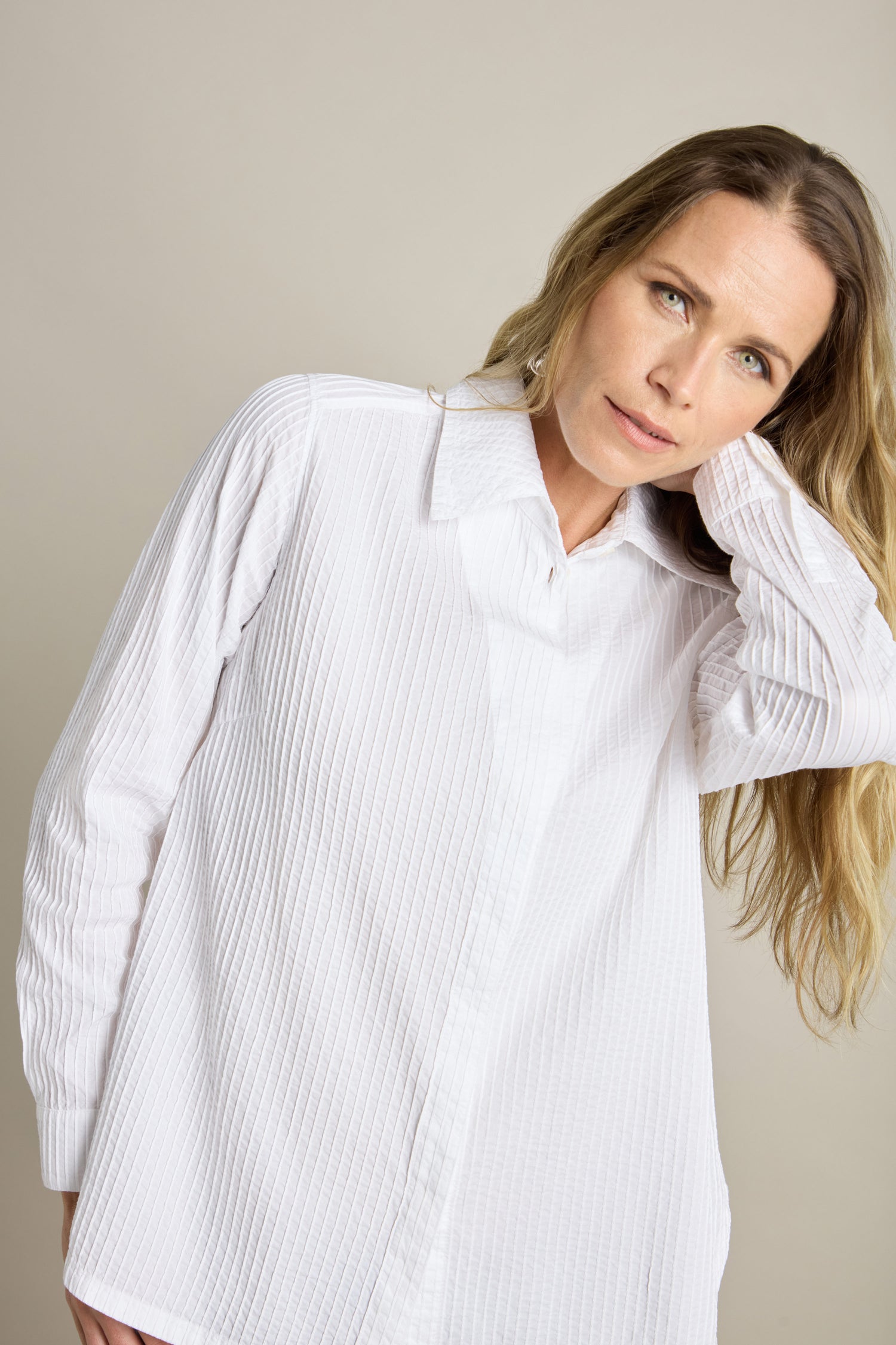 A person with long hair, wearing a textured white Fitted Pleat Shirt from Yacco Maricard, touches their head with one hand while standing against a plain background. The shirt enhances their tailored look.