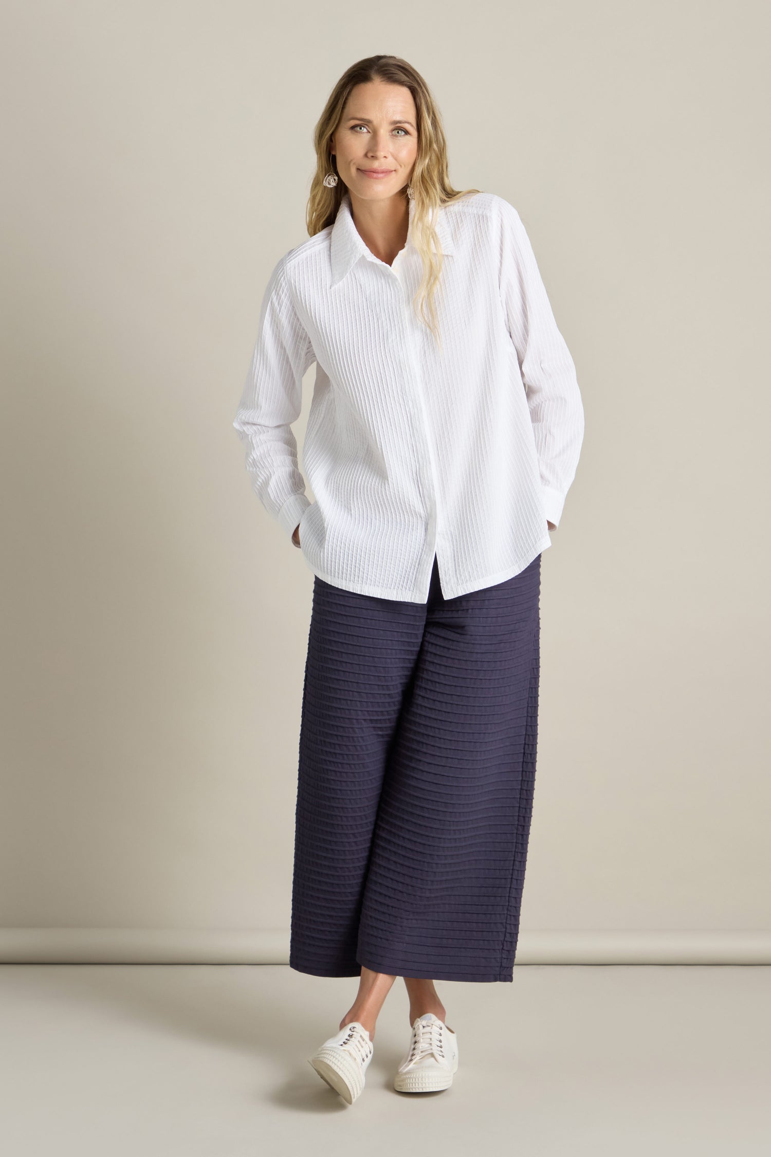 A woman stands against a plain background, wearing a Yacco Maricard Fitted Pleat Shirt, navy wide-leg pants, and white sneakers. She has her hands in her pockets and is smiling calmly at the camera.