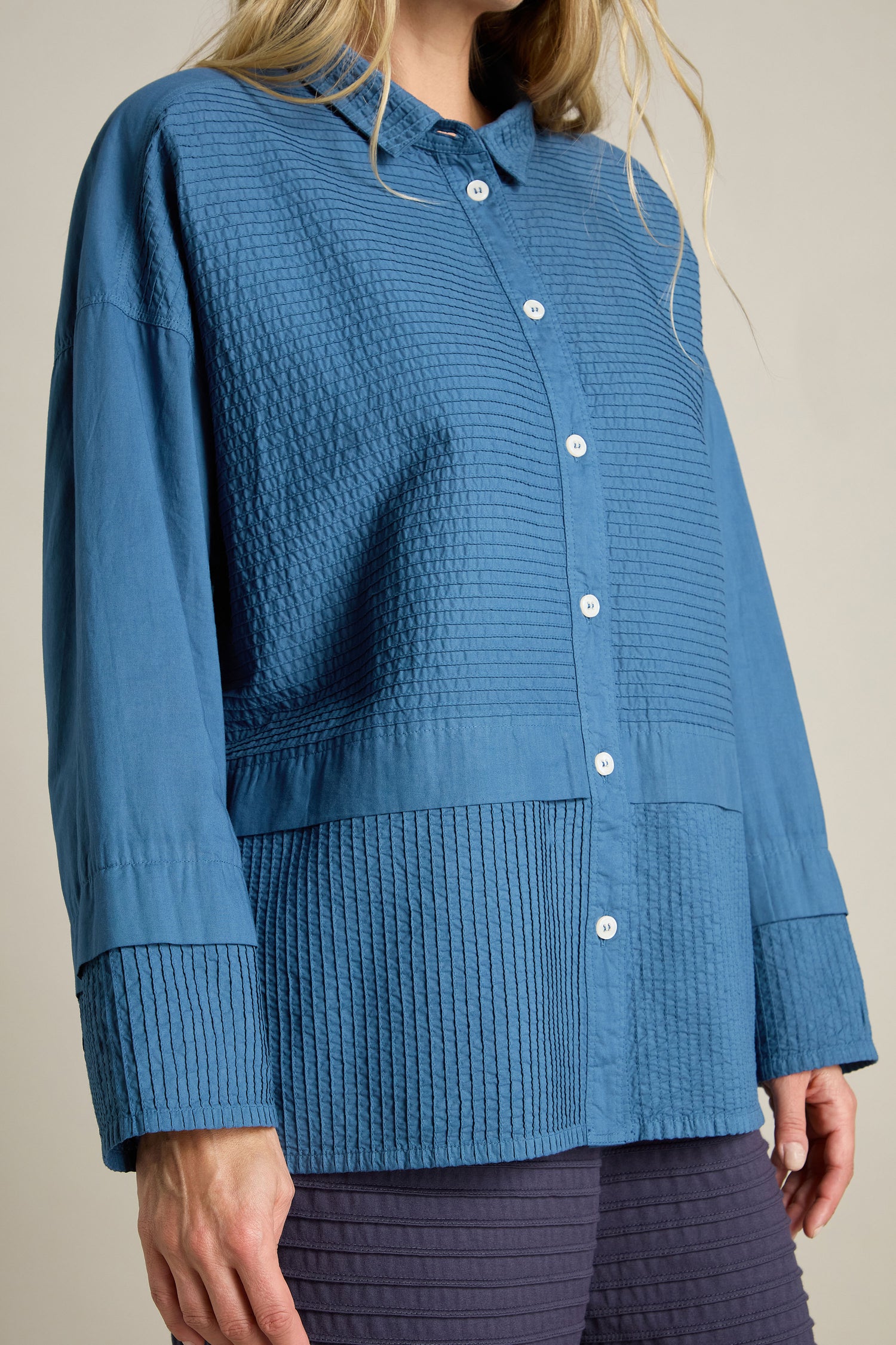 A person wearing a Deep Hem Pleated Shirt by Yacco Maricard, characterized by its blue textured fabric and long sleeves, standing against a neutral background.