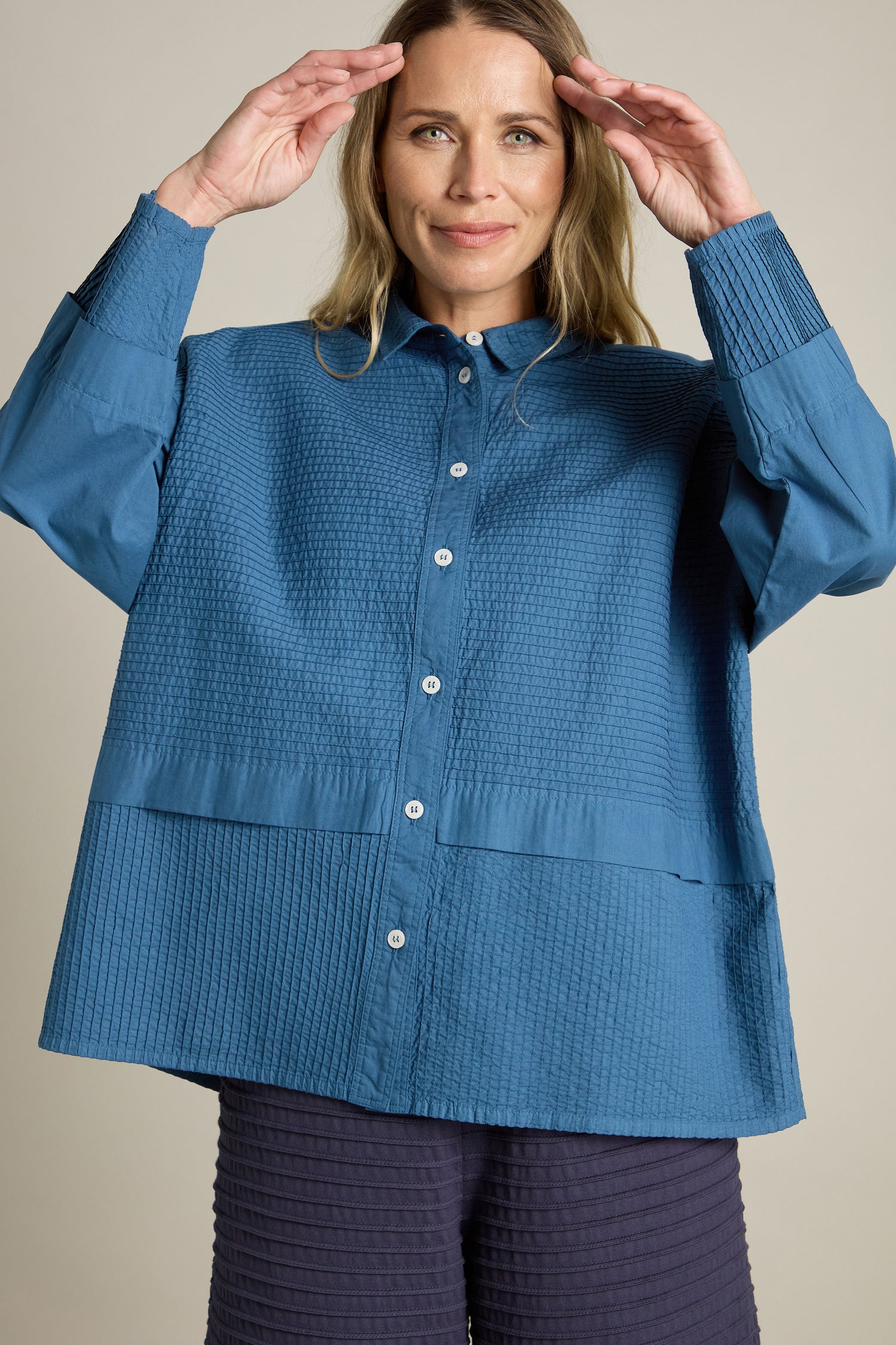 A person stands against a plain background wearing a Yacco Maricard Deep Hem Pleated textured blue shirt and dark pants, raising their hands near their head in a modern classic pose.