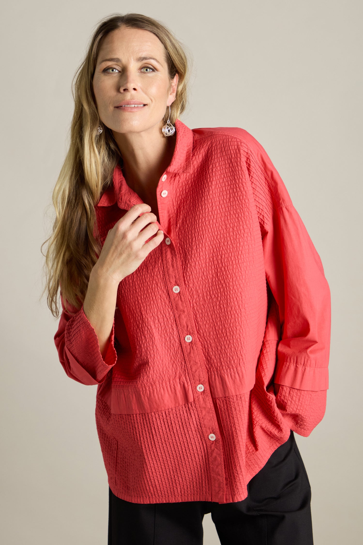 A woman with long blonde hair casually poses in a loose-fitting, textured Deep Hem Pleated Shirt by Yacco Maricard. She pairs the red shirt with black pants and stands against a plain background.