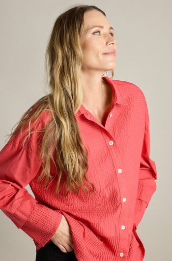 A woman with long, wavy hair is wearing a textured coral Deep Hem Pleated Shirt from Yacco Maricard. She strikes a pose with her hands on her hips against a plain background, gazing slightly upwards.