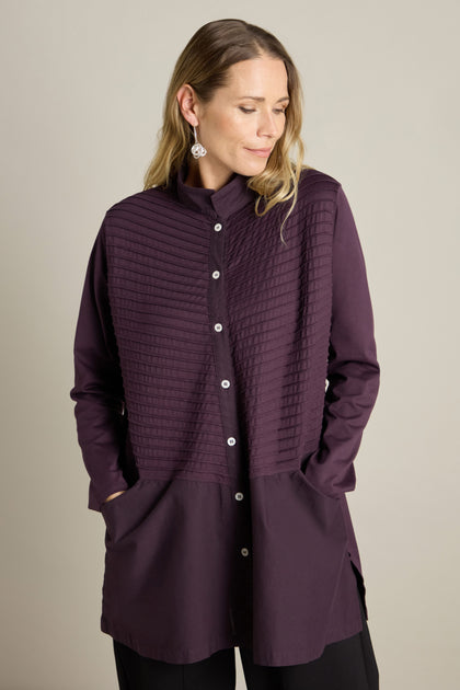 A woman stands against a plain background, wearing a long-sleeved, dark purple button-up Cotton Jersey And Calico Overshirt with horizontal ribbed detailing on the upper half and large pockets on the lower half.