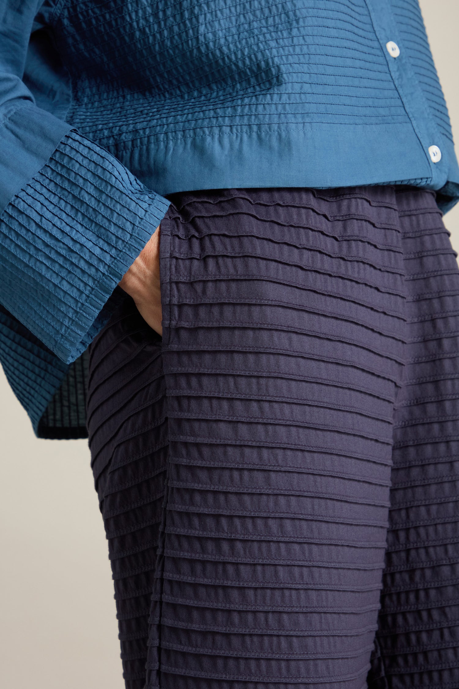 A close-up of a person wearing a textured blue shirt and Cotton Jersey Pleat Trousers by Yacco Maricard, with one hand in their pocket.