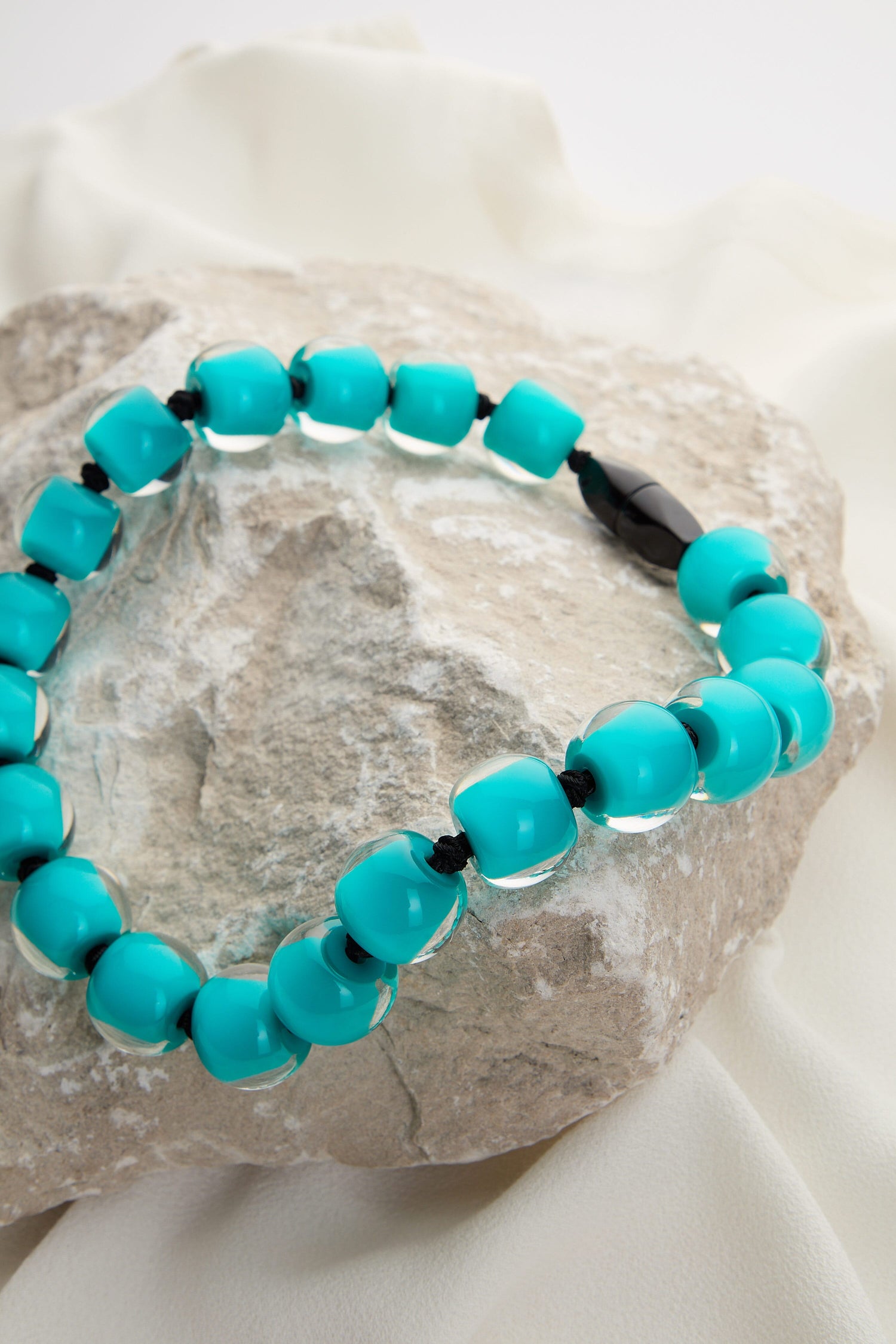 An Aqua Colourful Bead Necklace with vibrant blue resin beads and a single black bead is draped over a large, rough stone and rests on a white fabric background.