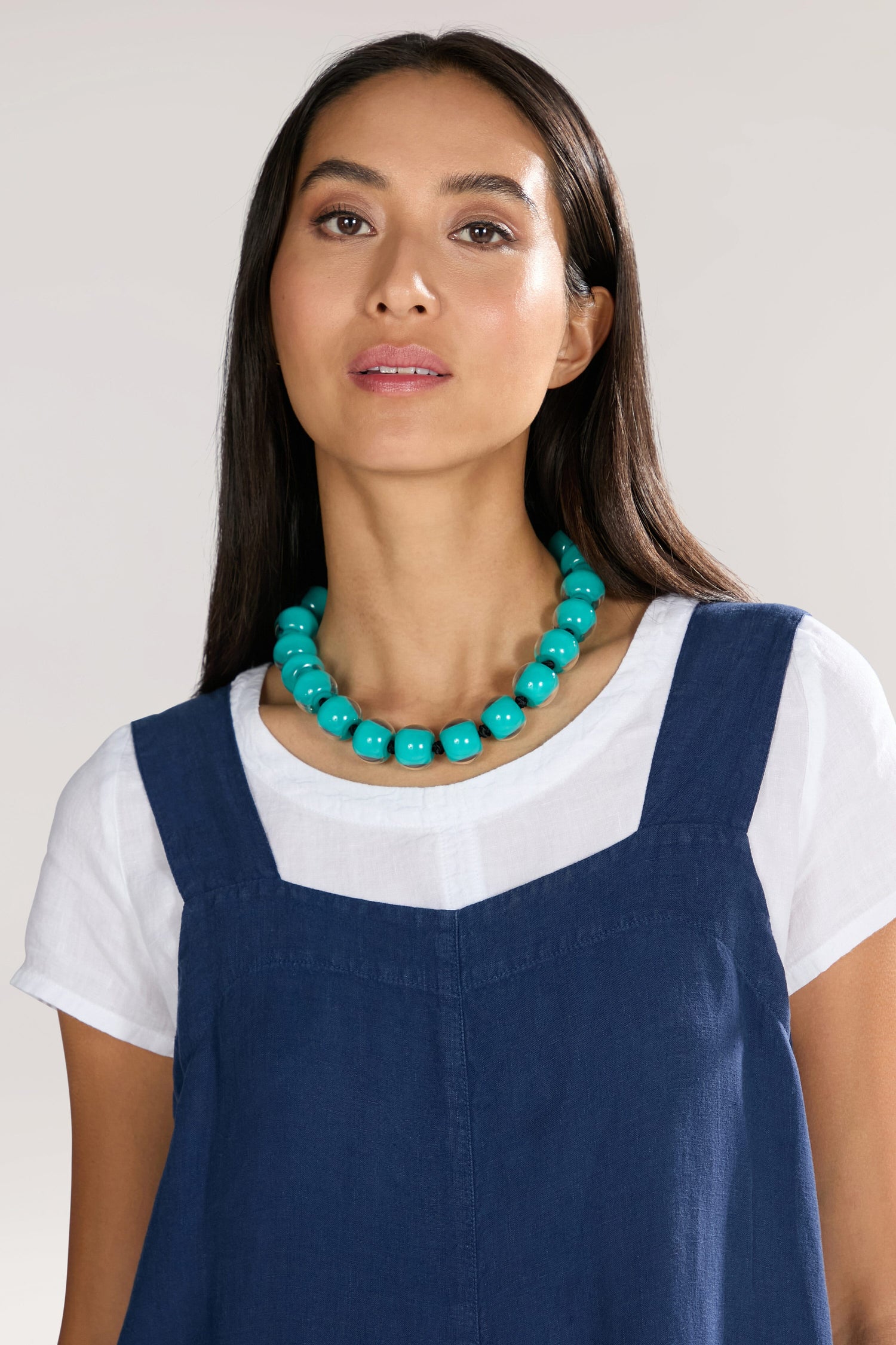 A person with long, dark hair is wearing a white top under a blue pinafore dress and an Aqua Colourful Bead Necklace featuring vibrant blue resin beads and a magnetic closure, looking directly at the camera against a plain background.