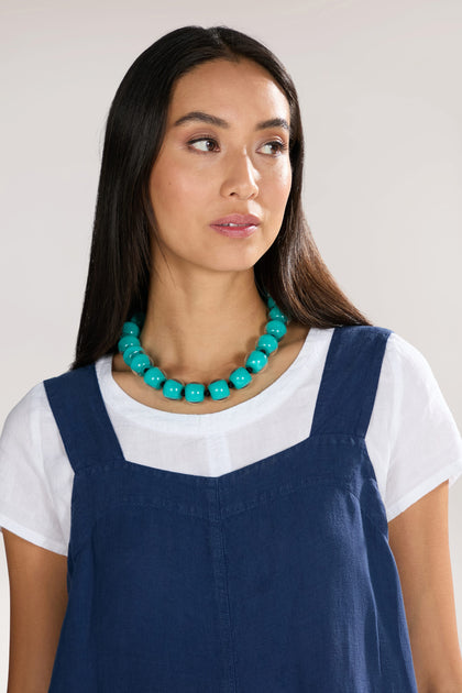 A woman with long dark hair wears a blue sleeveless dress over a white shirt, accessorized with an Aqua Colourful Bead Necklace, looking to her right.