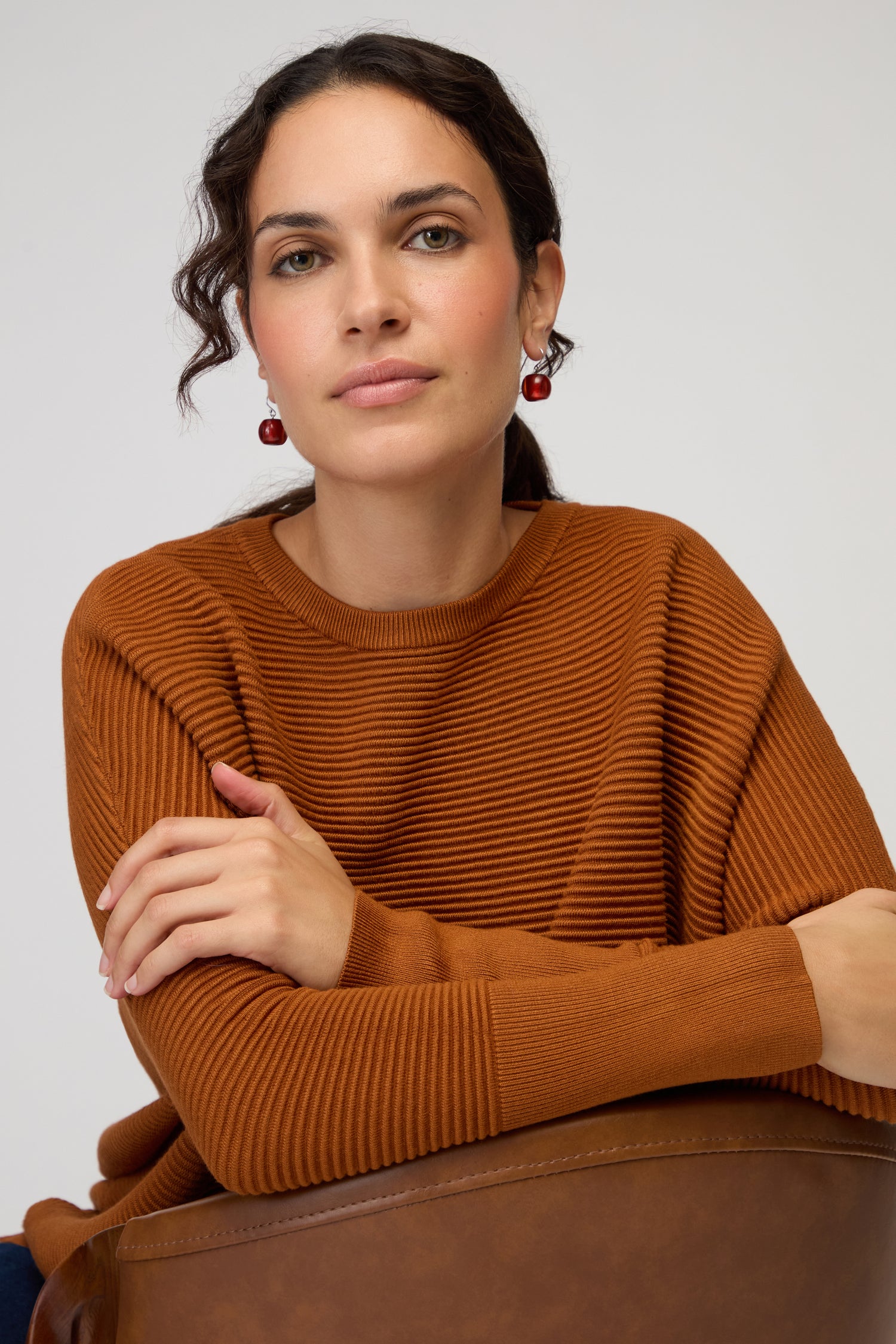 Woman with dark hair wearing a rust-colored ribbed sweater and Colourful Bead Resin Earrings, sitting with arms crossed and a brown bag in front of her, looking at the camera.