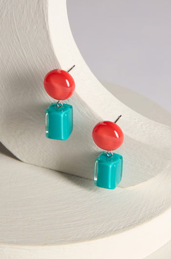 A pair of Sphere and Cube Resin Earrings featuring red circular tops and turquoise rectangular drops, elegantly displayed on a curved, white backdrop.