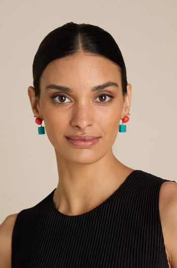 A woman with dark hair tied back, wearing statement red and green Sphere and Cube Resin Earrings and a sleeveless black top, looking directly at the camera.