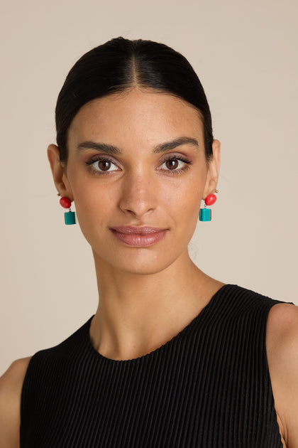 A woman with dark hair tied back, wearing statement red and green Sphere and Cube Resin Earrings and a sleeveless black top, looking directly at the camera.