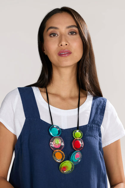 A woman with long dark hair, wearing a white shirt under a blue sleeveless dress, complements her look with a vibrant Resin Flower Necklace.