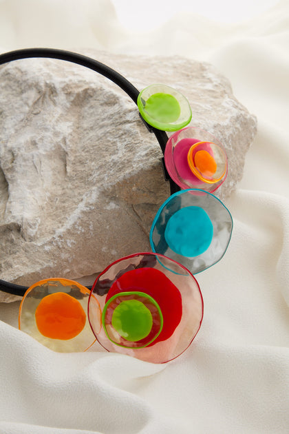 A Short Resin Flower Necklace with circular, transparent decorations in green, pink, blue, orange, and red rests on a textured rock against a white fabric background—a true piece of artistic expression.