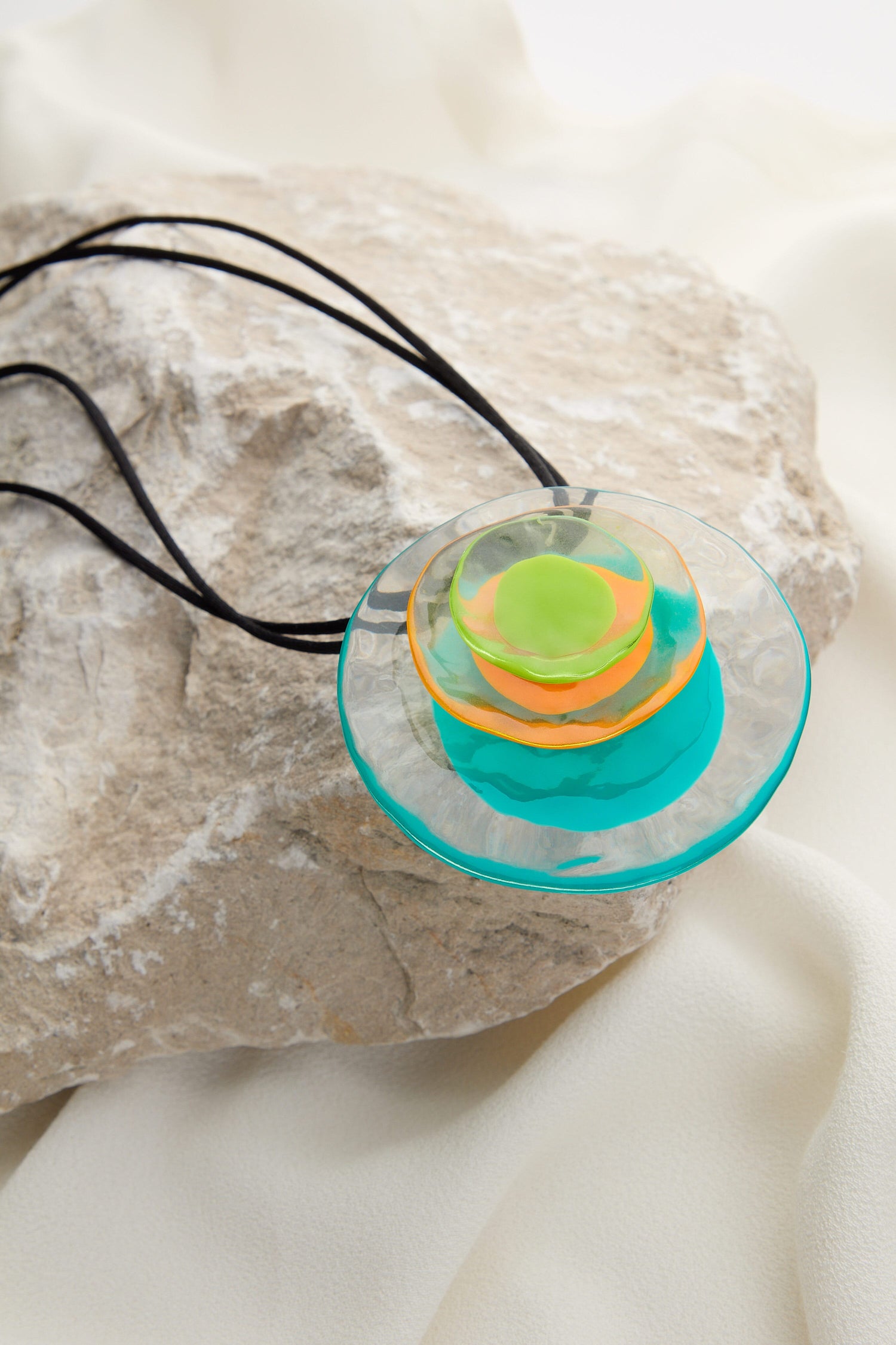 A Resin Flower Pendant with concentric layers in green, orange, and blue on a black cord, rests on a gray stone background. The cord offers an adjustable length for a perfect fit.