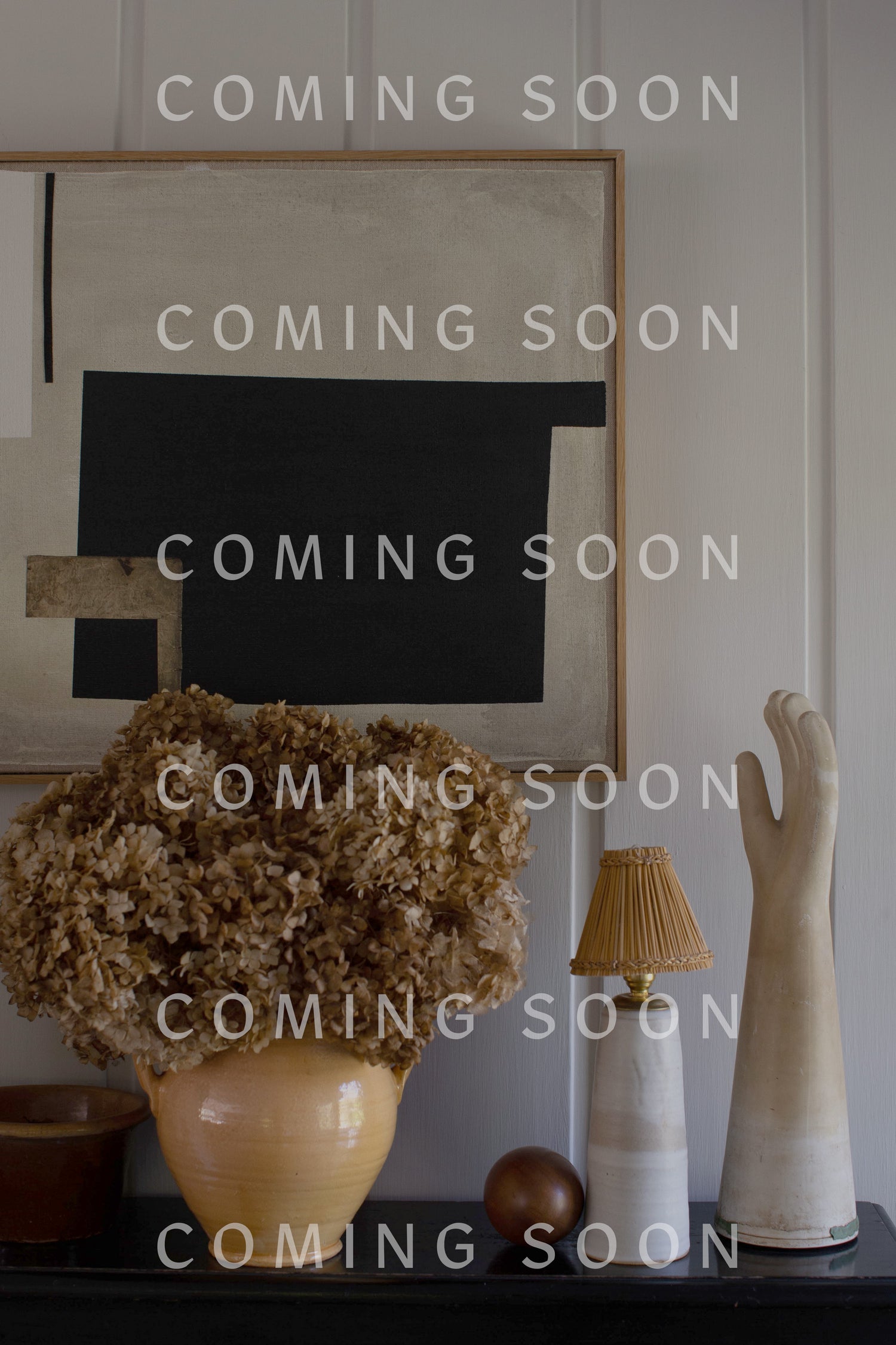 A beige vase with dried flowers, a lamp, a decorative hand, and a wooden ball are arranged on a surface in front of a black and beige abstract painting. The scene exudes the same elegance as the Cashmere Blend Crop Jumper. "COMING SOON" text is repeated across the image.