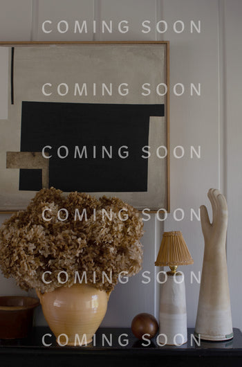 A beige vase with dried flowers, a lamp, a decorative hand, and a wooden ball are arranged on a surface in front of a black and beige abstract painting. The scene exudes the same elegance as the Cashmere Blend Crop Jumper. "COMING SOON" text is repeated across the image.