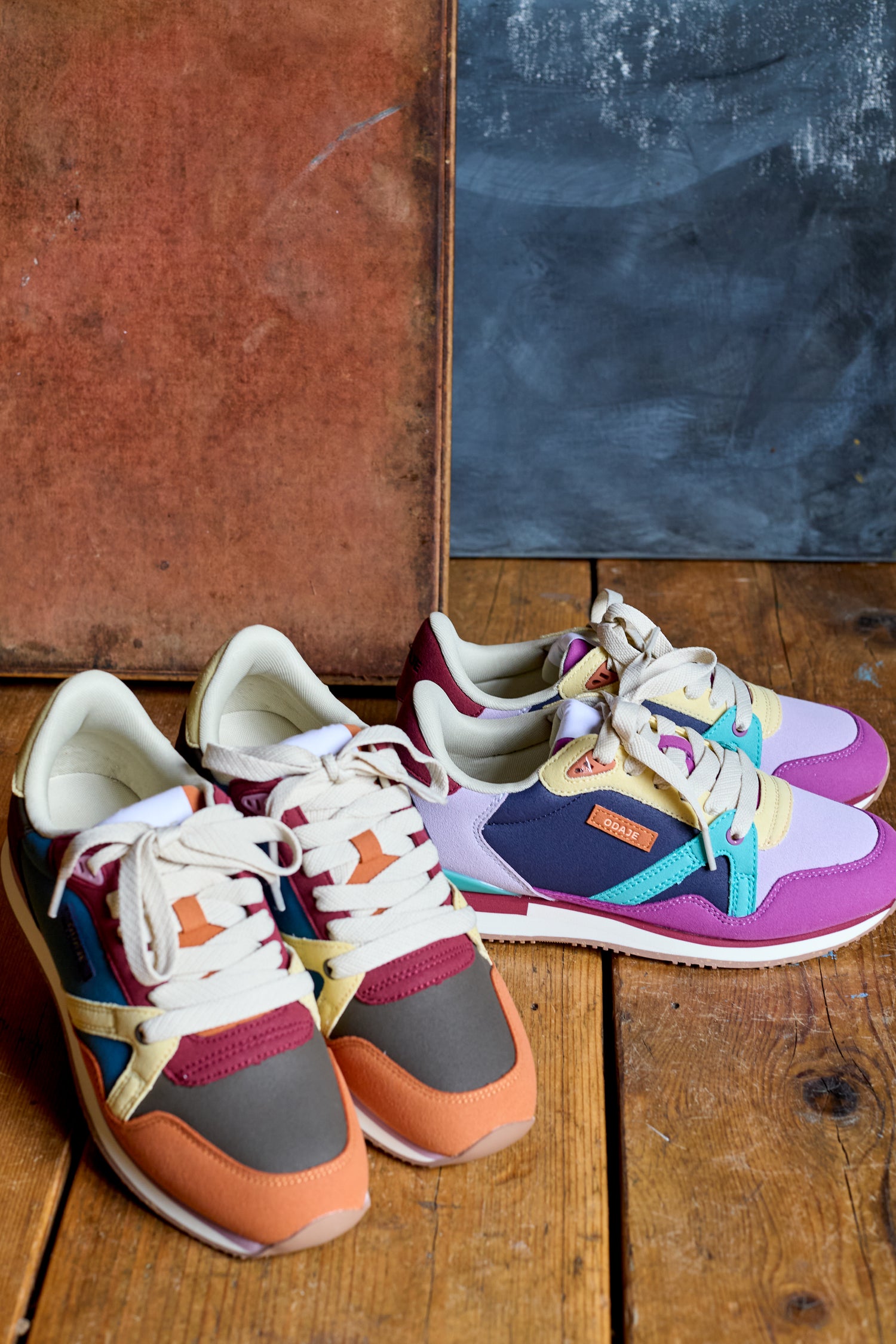 Two pairs of Colour Pop Andree Trainers are placed side by side on a wooden floor, set against a textured backdrop with brown and blue panels.