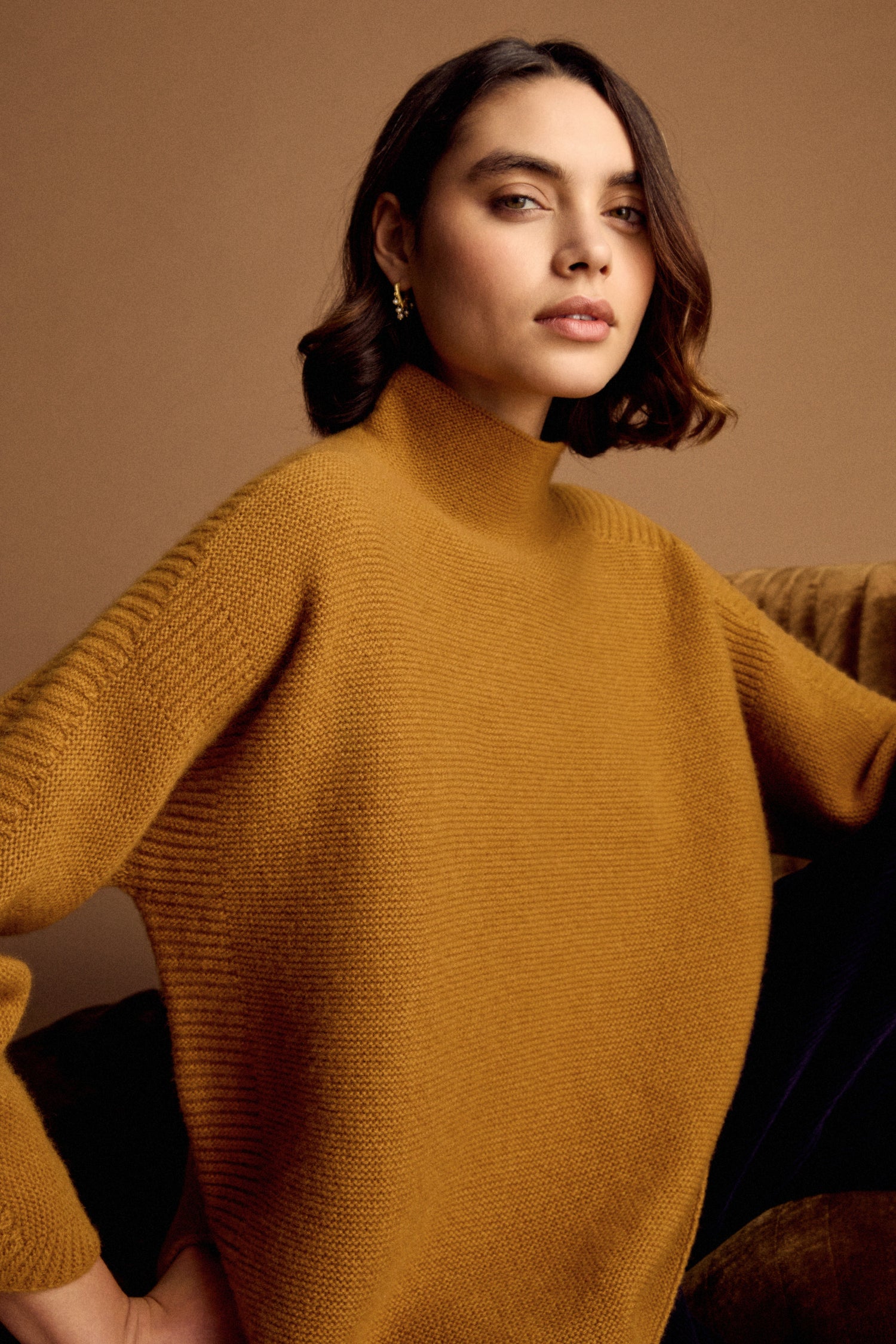 A woman with short brown hair wears the Merino Wool Stitch Detail Jumper in mustard, sitting on a brown couch against a beige background, highlighting its relaxed boxy silhouette—an ideal knitwear staple.